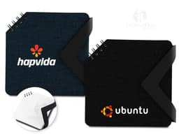Mini caderno preto de couro aberto sobre uma mesa de escritório, ao lado de um notebook e diversos dispositivos eletrônicos.