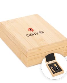 Kit vinho 2 peças em uma caixa de papelão marrom. A embalagem é de carton corrugado e contém um objeto de madeira em forma de caixa de transporte. A caixa está fechada e possui detalhes em textura de madeira. No fundo da imagem, há um bloco de madeira e vários livros empilhados.