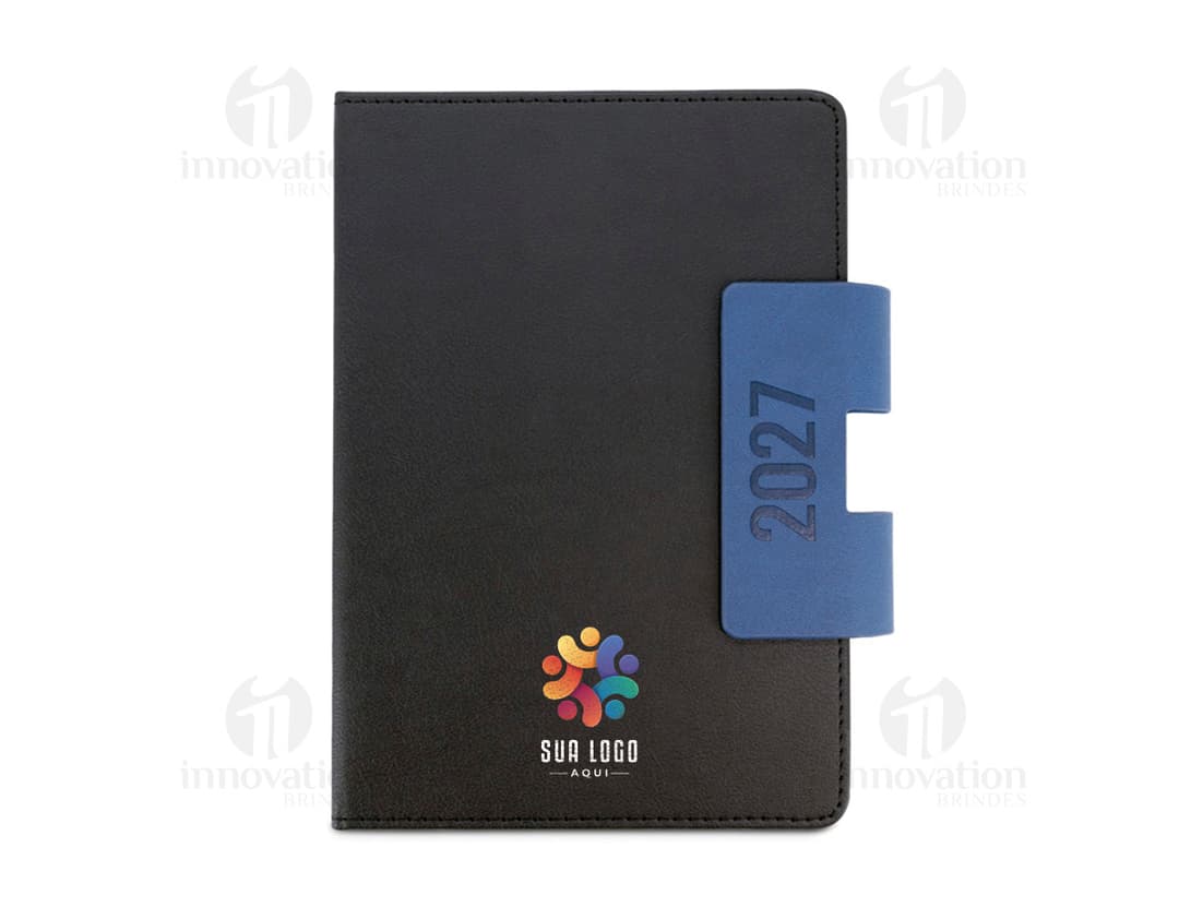Notebook preto aberto, com teclado e tela exibindo ícones e botões. Ao lado, uma agenda A5 com capa dura em PU, destacando-se